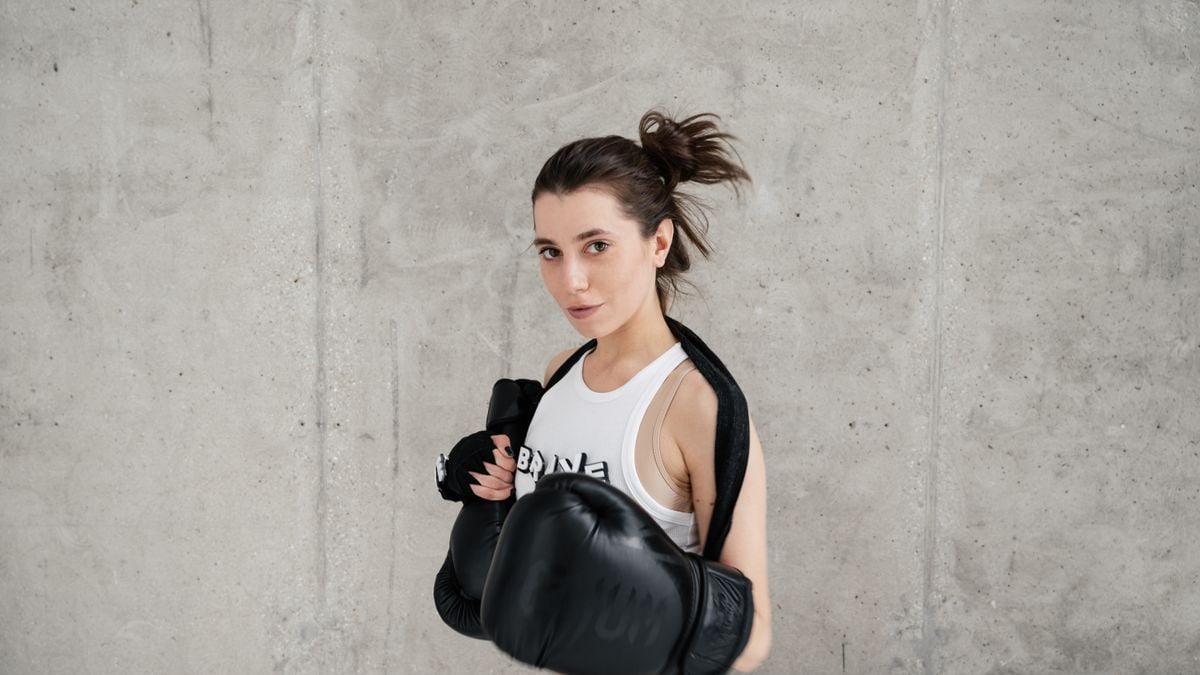 Confident young woman posing with boxing gloves against a concrete wall, showcasing style and sportiness.