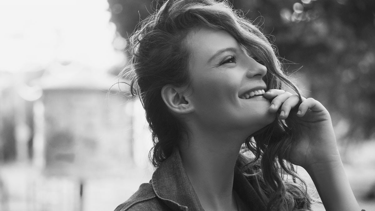 Black and white side view of smiling female with hand at chin and curly hair covering face face looking into distance while standing on street on blurred background
